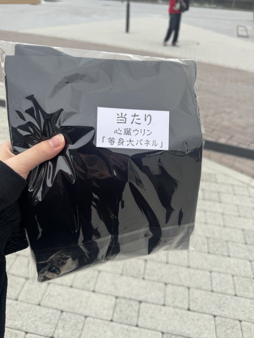 A hand holds a black plastic bag with a label in Japanese reading '当たり 心臓くりん「等身大パネル」' indicating a winning prize of a life-size panel.