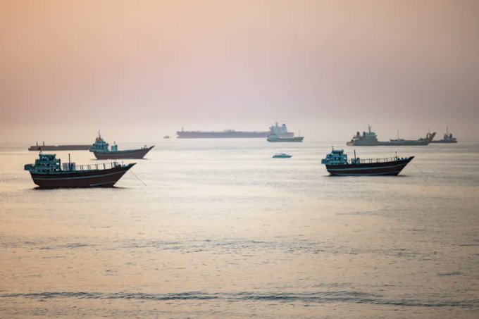Several cargo and oil tanker ships sit anchored in the calm waters of the Gulf of Oman at dusk, illustrating the maritime activity where TankerTrackers.com documented at least 19 ship-to-ship transfers of oil and energy products.