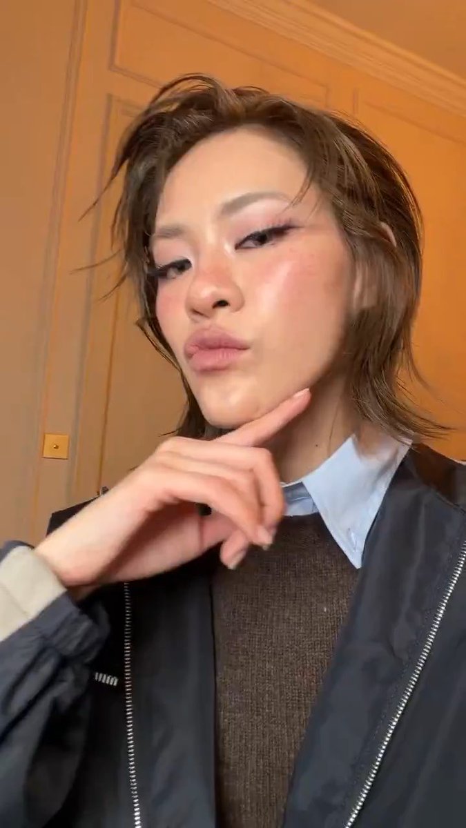A close-up selfie of a young woman with brown hair wearing a dark jacket over a sweater and blue collared shirt, posing with her hand near her chin.