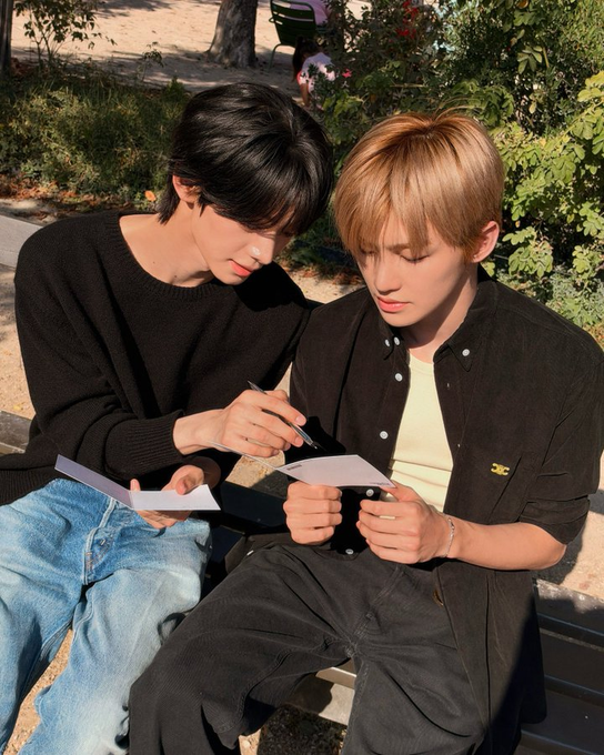 Two young men sit together outdoors, one writing in a notebook while both look down at papers in their hands.