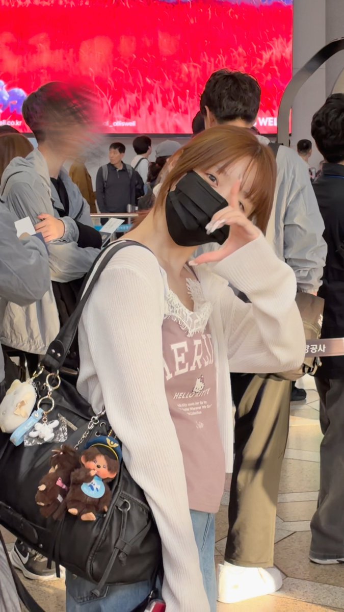 A young woman wearing a black mask poses in a crowded venue, with visible text on her shirt reading 'AER', 'HELLO KITTY', and 'Whoever We Go'.
