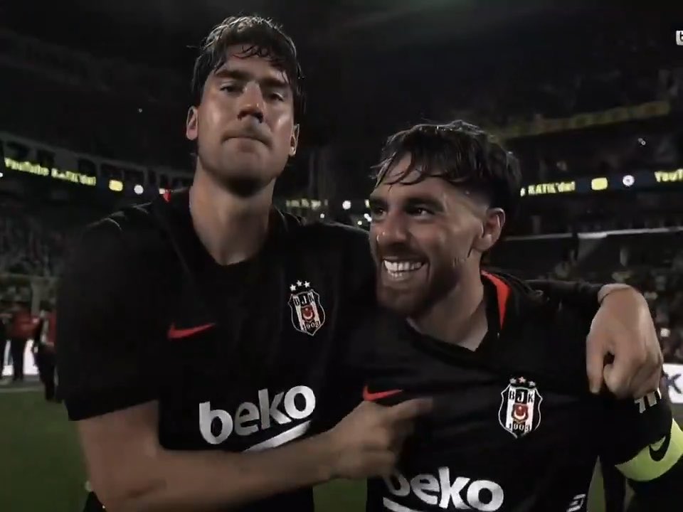 Two Beşiktaş football players in black kits with the club crest embrace and celebrate after a match, with one wearing a captain's armband, in a stadium setting.