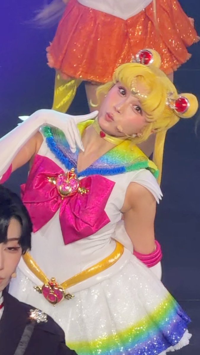 A performer on stage dressed as Super Sailor Moon, wearing a white bodice with a large pink bow, rainbow-colored collar and skirt accents, and a blonde wig with the iconic odango hair buns.