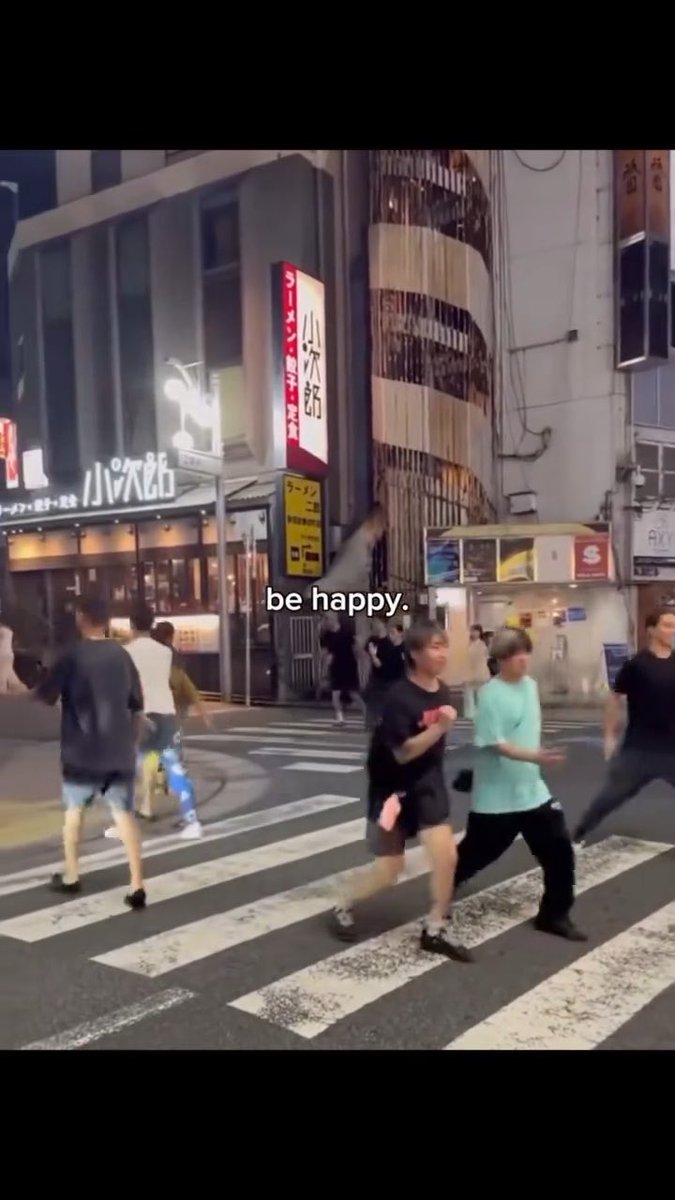 The image captures a lively night scene in Japan, featuring pedestrians crossing a crosswalk in front of storefronts with Japanese signage, including "ハクション大魔王" (Hakushon Daimao) and "ラーメン" (Ramen). The caption "be happy." overlays the bustling street, complementing the energetic urban atmosphere as people casually stroll through the vibrant cityscape.
