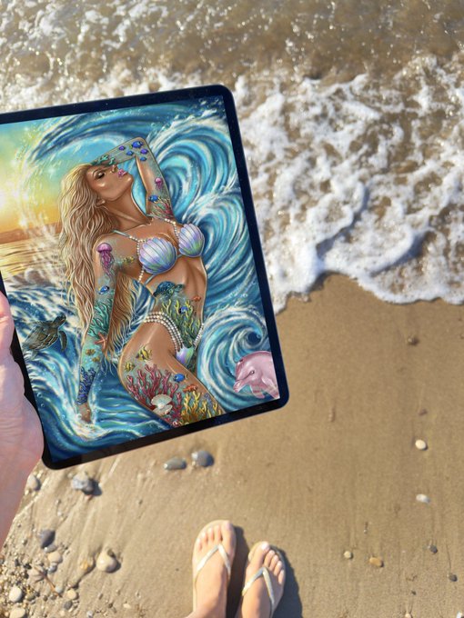 A hand holds a tablet displaying an illustration of a blonde mermaid in ocean waves, set against the backdrop of sandy beach and incoming surf with feet in flip-flops visible below.