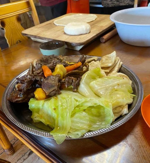 A plate of slow-cooked meat with carrots, cabbage, and flatbread on a metal platter, with a wooden cutting board, dough, and a white bowl in the background.