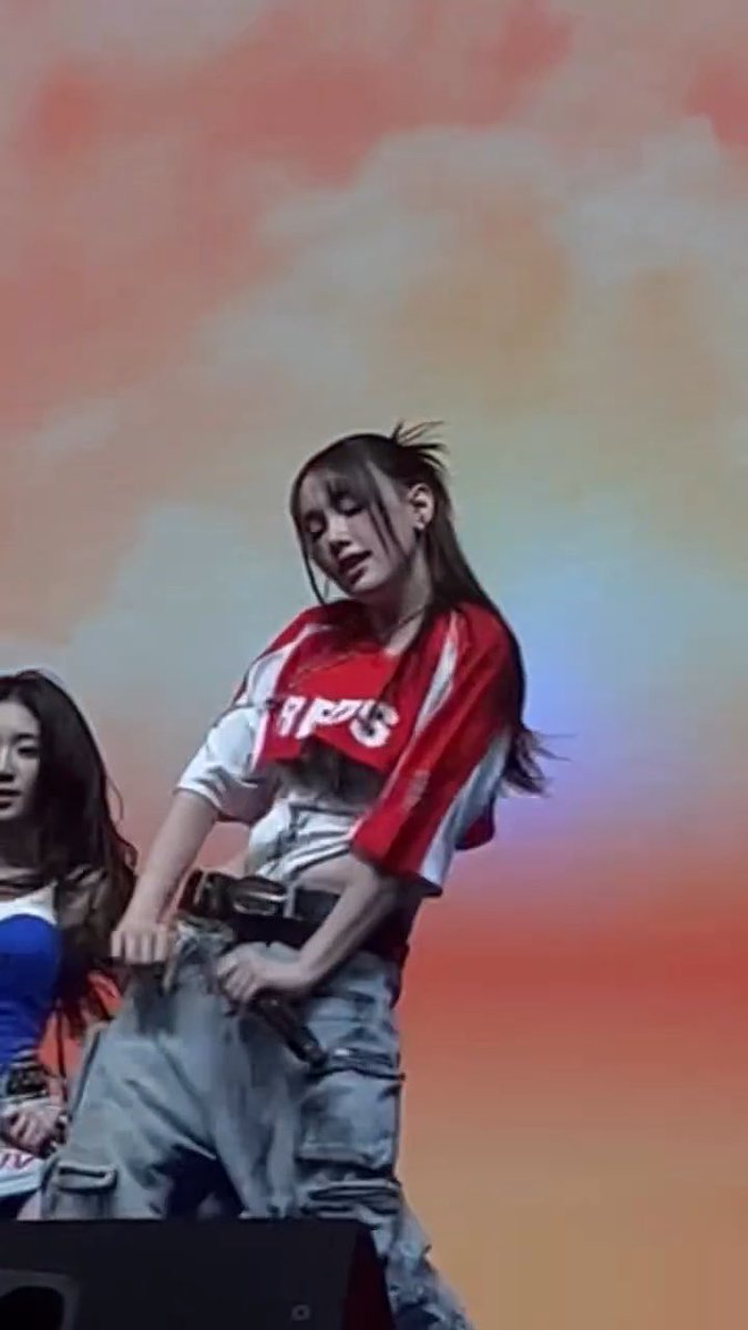 A young woman performs on stage wearing a red and white top with a harness, against a colorful pink and blue background.