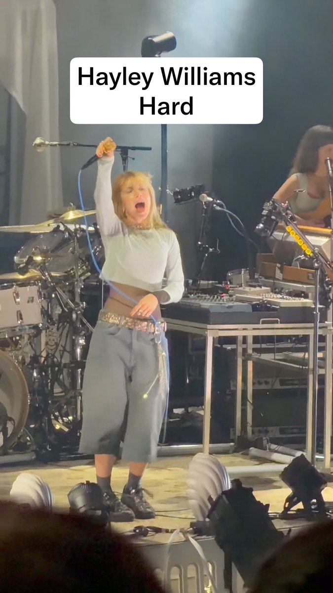 A concert photo captioned 'Hayley Williams Hard' showing the Paramore singer performing energetically on stage, wearing a white long-sleeved crop top and blue pants with a bedazzled belt.