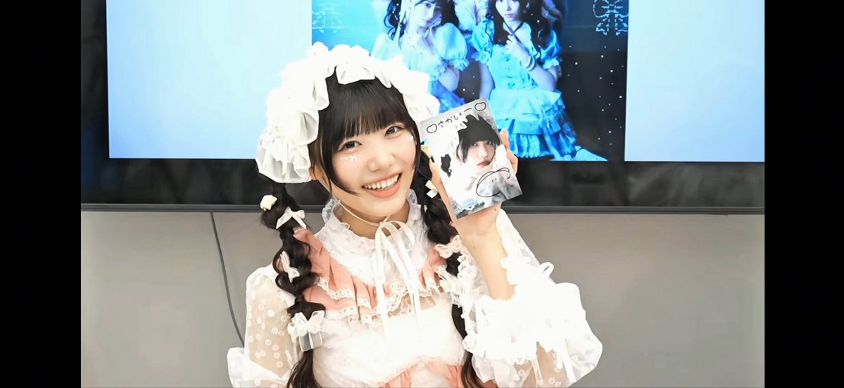 A young woman in a pink and white frilly lolita outfit with twin braids smiles while holding up a signed card or photobook, with a screen displaying blue imagery behind her.