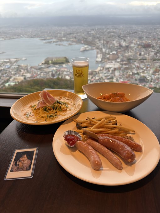 A meal of sausages, fries, salad, and kimchi with a glass of 'Classic' beer sits on a table overlooking a coastal city view, with a small photograph resting beside the plate.