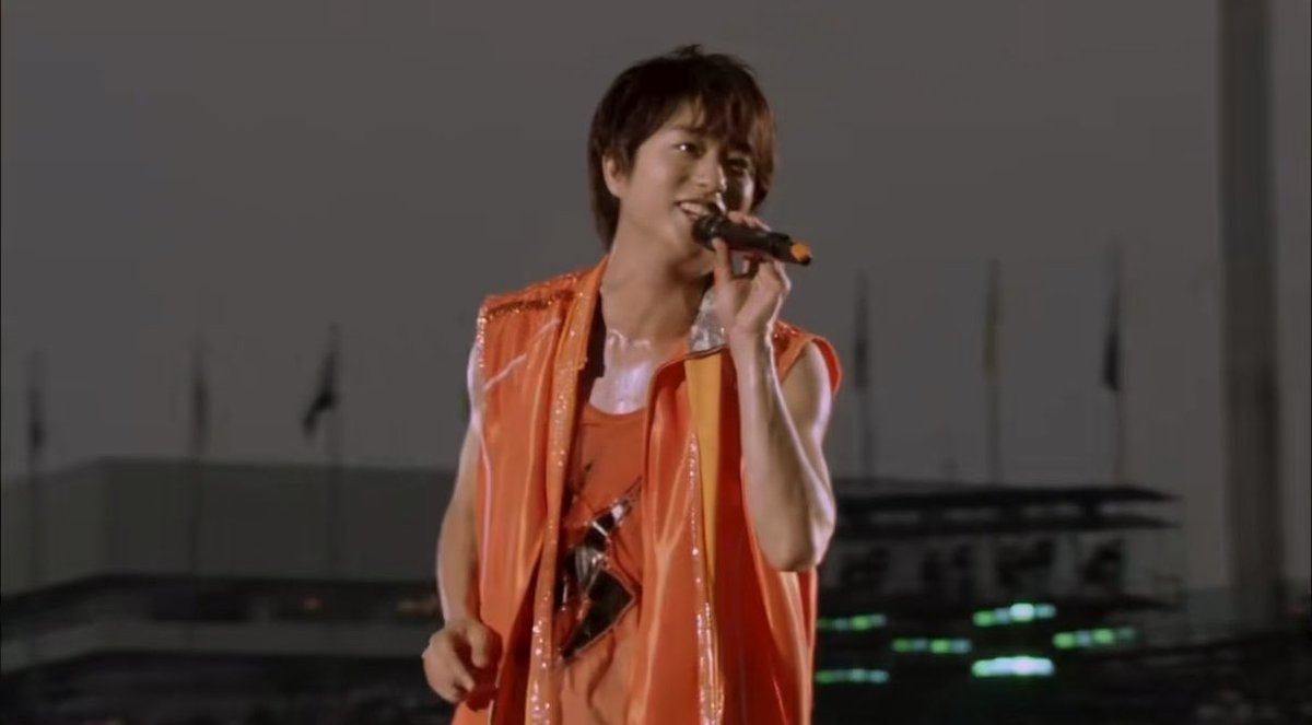 A young man in an orange vest sings into a microphone on a rooftop stage, smiling as he performs. The accompanying text humorously exaggerates the "destructive power" of his performance, joking about his neck snapping and the need to declare a state of emergency.
