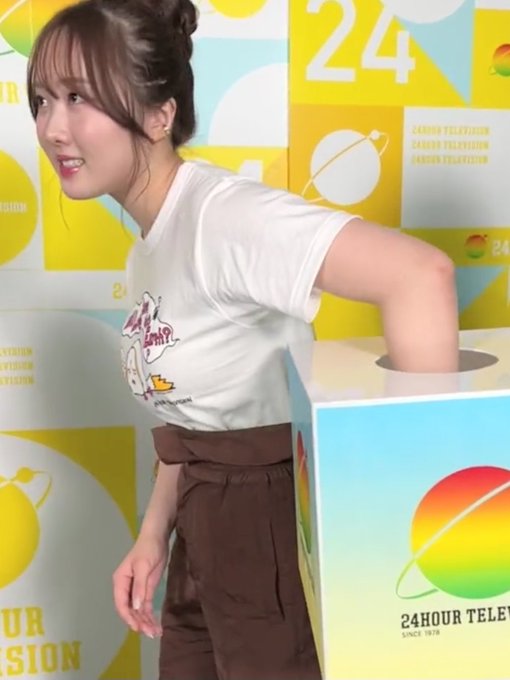 A young Japanese woman wearing a white graphic t-shirt and brown skirt posing in front of a yellow '24HOUR TELEVISION SINCE 1978' backdrop, with her arm reaching into a box.