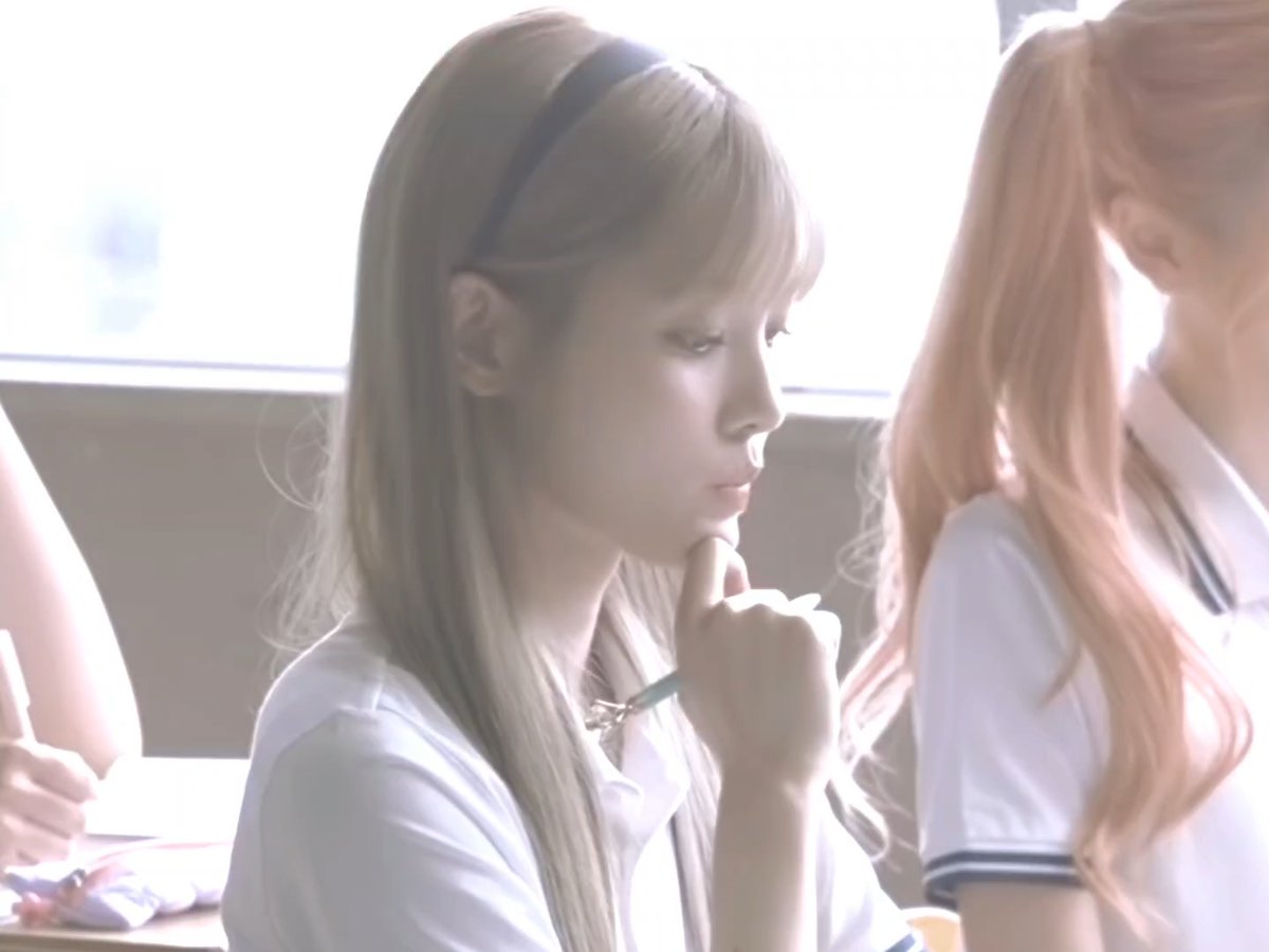 A young woman with long dark hair wearing a black headband and white shirt, looking to the side in a brightly lit room with other people partially visible.