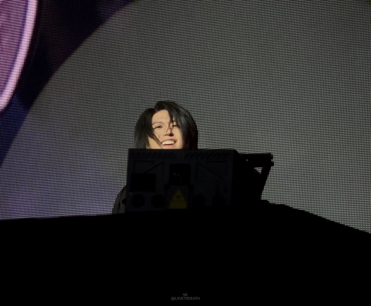 A concert scene with a performer with dark hair smiling behind DJ/laptop equipment, with a large pixelated LED screen in the background and the watermark '@ILXVETODEATH' at the bottom.