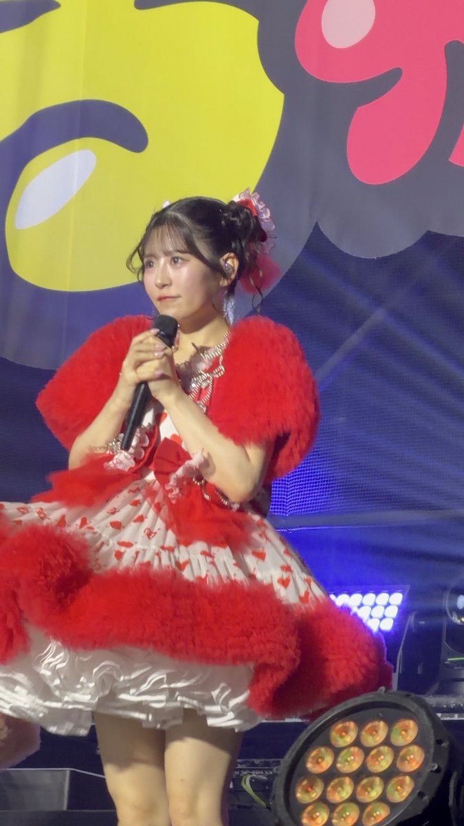 A young woman in a red and white frilly dress with feathered shoulders holds a microphone on stage during what appears to be an idol concert.