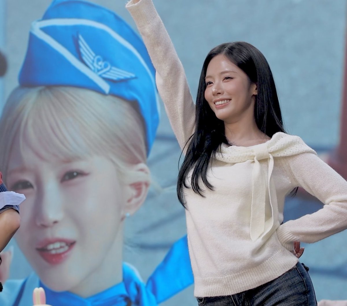 A smiling East Asian woman with long black hair, wearing a cream-colored sweater with a bow, raises her arm in celebration while standing in front of a large screen displaying an idol in a blue flight attendant outfit.