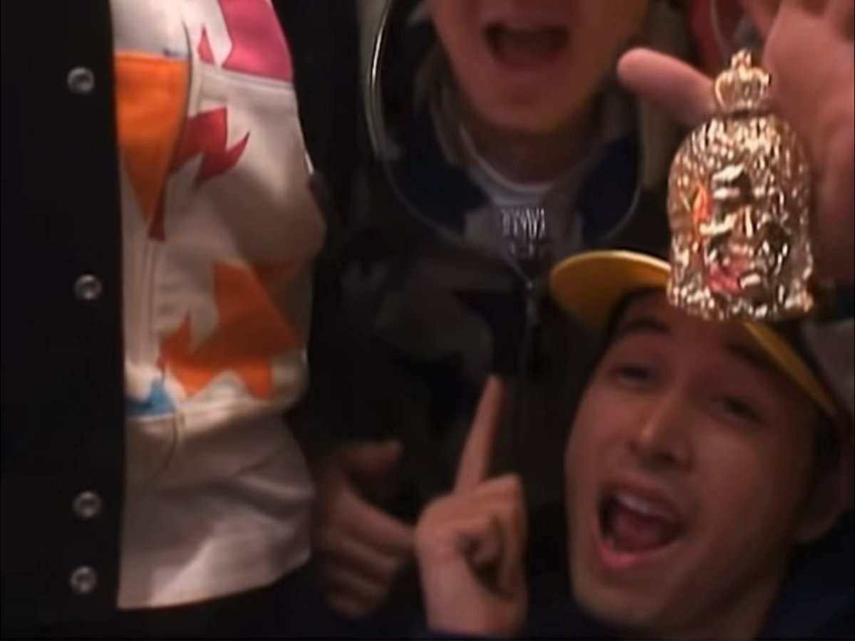 A close-up of excited people indoors, with one person wearing a yellow cap featuring a gold pineapple ornament.
