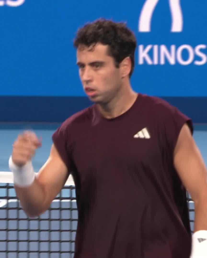 Jaume Munar, wearing a maroon Adidas sleeveless shirt, clenches his fist in a determined gesture on the tennis court, with a "KINOS" sponsor banner visible on the blue backdrop behind him, capturing his gritty comeback to win the first set 7-5 over Carlos Alcaraz at Tokyo.