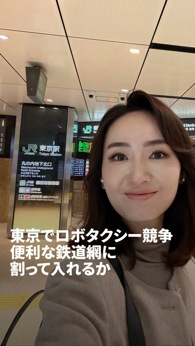 A woman takes a selfie at Tokyo Station, with Japanese text overlay reading "東京でロボタクシー競争 便利な鉄道網に 割って入れるか" (Can robot taxis break into Tokyo's convenient rail network?).