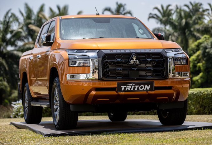 An orange Mitsubishi 'THE NEW TRITON' pickup truck displayed on a platform with tropical foliage in the background.