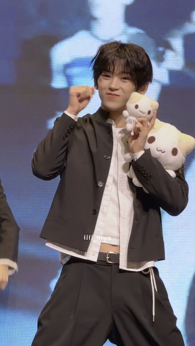 A young man with dark hair, identified by the post as Kim Dohun of IDNTT (아이덴티티), strikes a playful pose on stage with one fist raised while holding cute plush toys. He wears a black jacket over a white shirt paired with black pants, matching the caption's fun "Meow meow" vibe with the adorable cat-like stuffed animals he's holding up to the camera.
