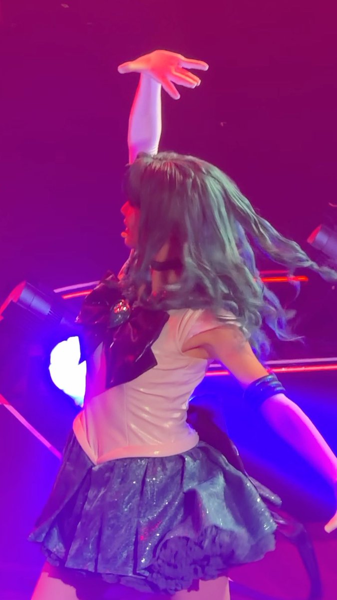 A performer with long teal hair, wearing a sailor-style school uniform with a black bow and blue skirt, striking a dynamic pose with arm raised on a stage lit in vibrant pink and blue lighting.