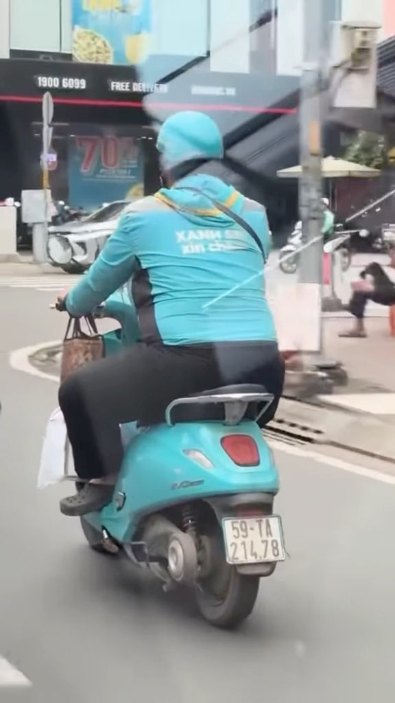 A Grab/Xanh SM delivery rider on a teal scooter with license plate '59-TA 214.78' riding down a city street, carrying a paper bag on the handlebars.