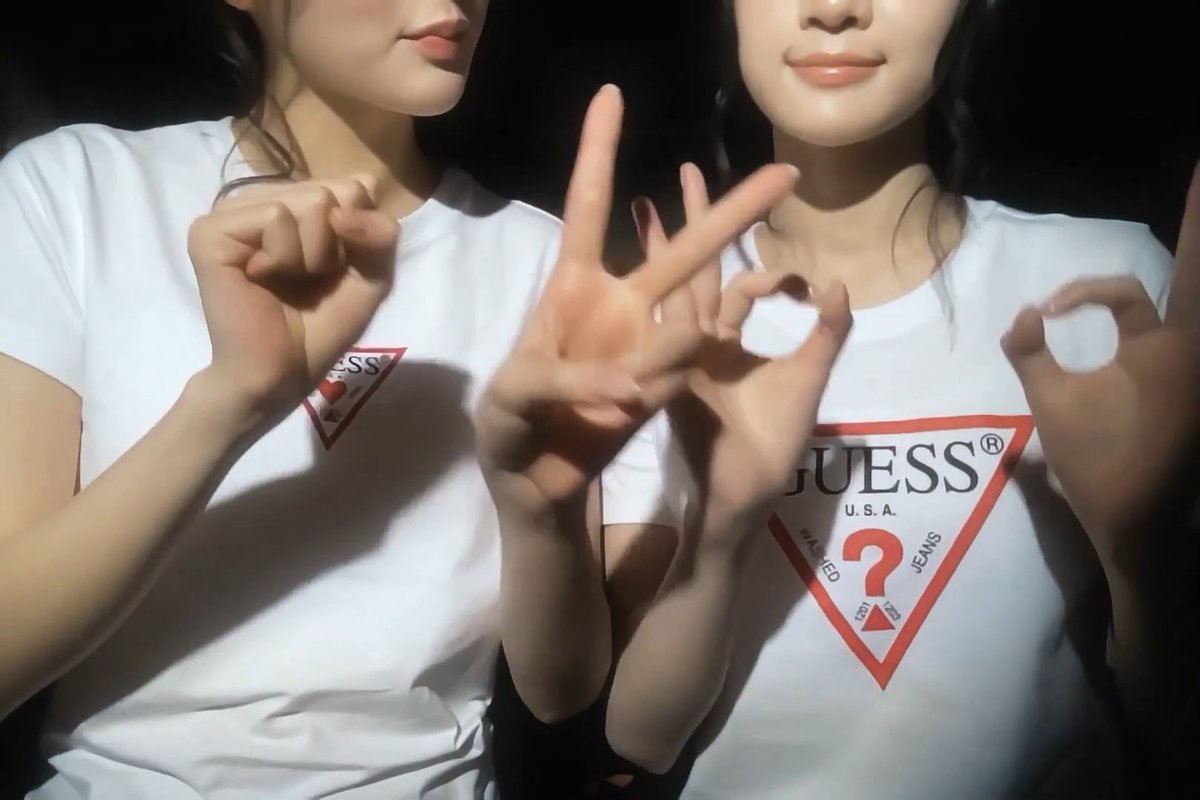 Two young women wearing matching white Guess U.S.A. t-shirts with the red triangle logo posing with hand gestures in a dark setting.