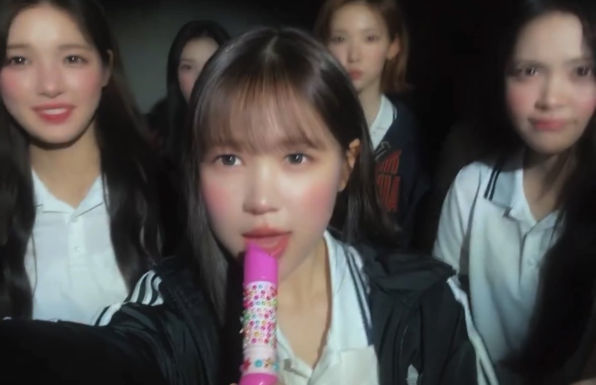 A group of young women wearing school-style uniforms, with the central figure holding a pink toy microphone.