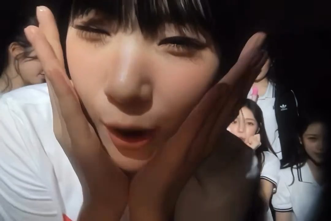 Close-up selfie of a person with their face framed by both hands, showing other individuals in white shirts in the blurred background.