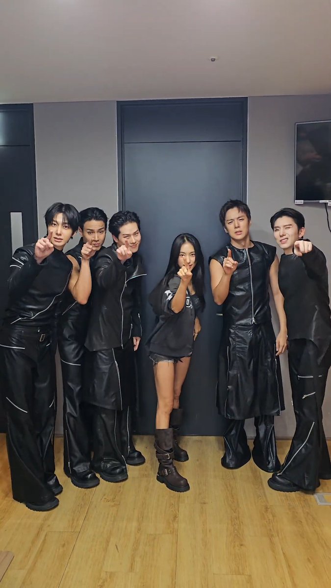 A group of six people posing together, with a woman in the center wearing an oversized black t-shirt and shorts, flanked by five men dressed in black leather outfits, all pointing at the camera.