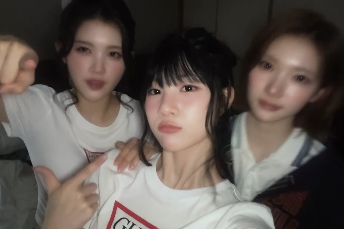 Three young women in white shirts take a close selfie together, with the center girl making a peace sign and looking at the camera.