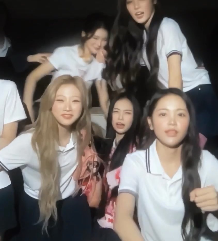 A group of young women, some in white school uniform shirts, pose closely together in what appears to be a dimly lit setting.