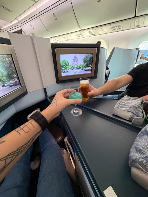 A first-person view inside an airplane cabin where two passengers are clinking glasses of drinks in a toast, with a cityscape visible on the seatback screen.