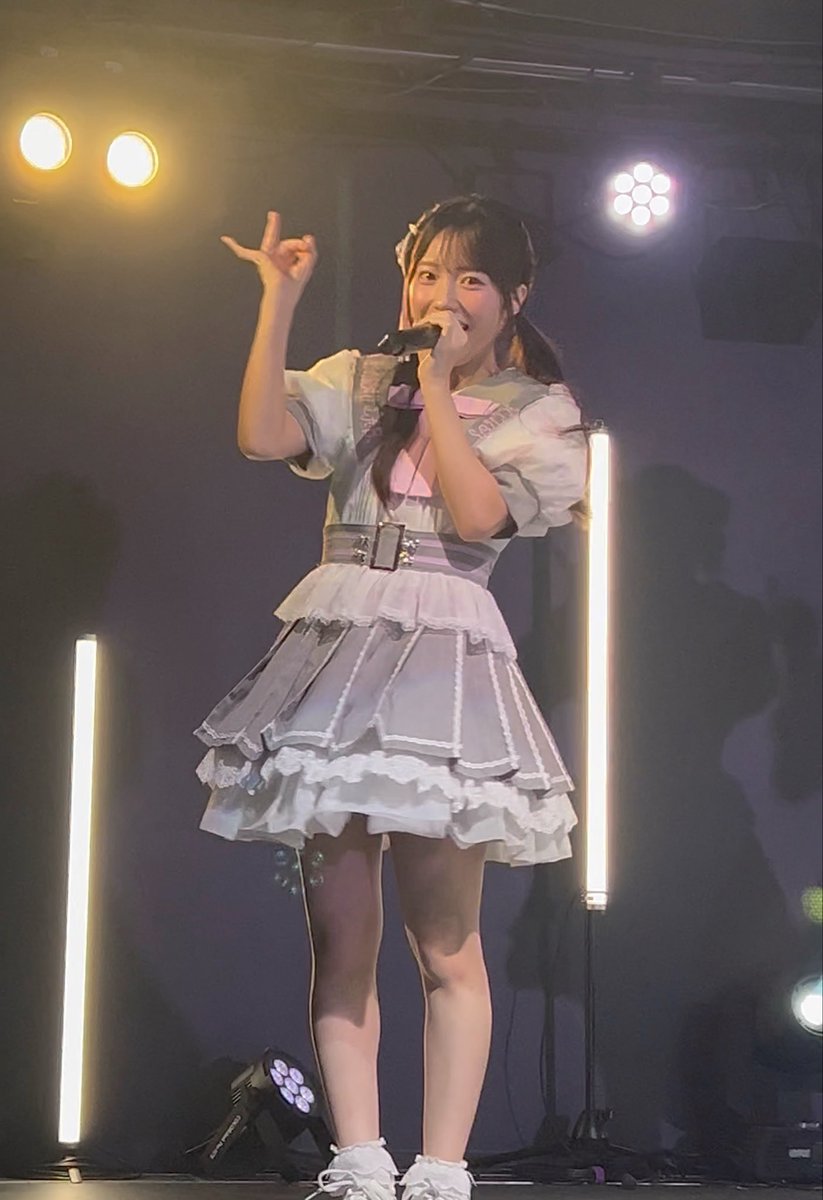 A young woman in a gray and white frilly idol costume sings into a microphone on stage, posing with one hand raised.