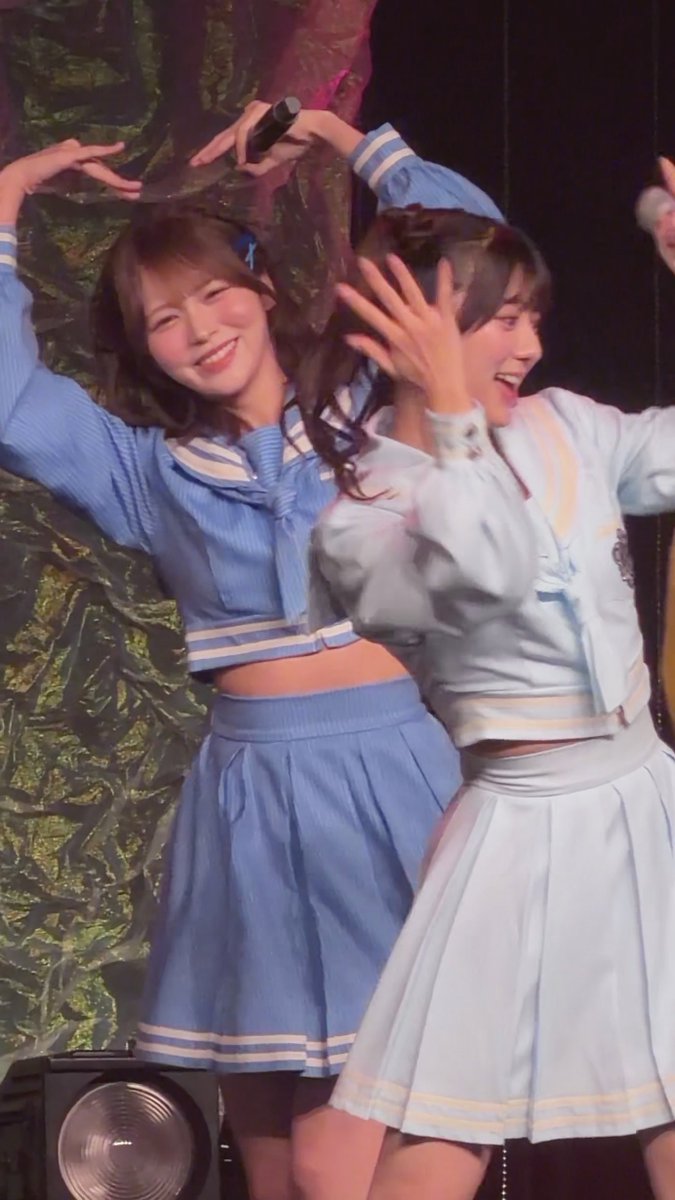 Two young women in sailor-style school idol outfits (one blue, one light blue/white) perform on stage, smiling and making heart shapes with their arms.