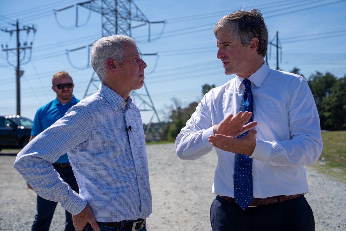 Two men talk outdoors near electrical transmission infrastructure, while another man stands behind them. There is no prominent text.