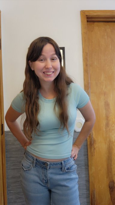 A woman with long wavy hair, a nose ring, and a light blue ribbed crop top standing and smiling in front of a wooden door.