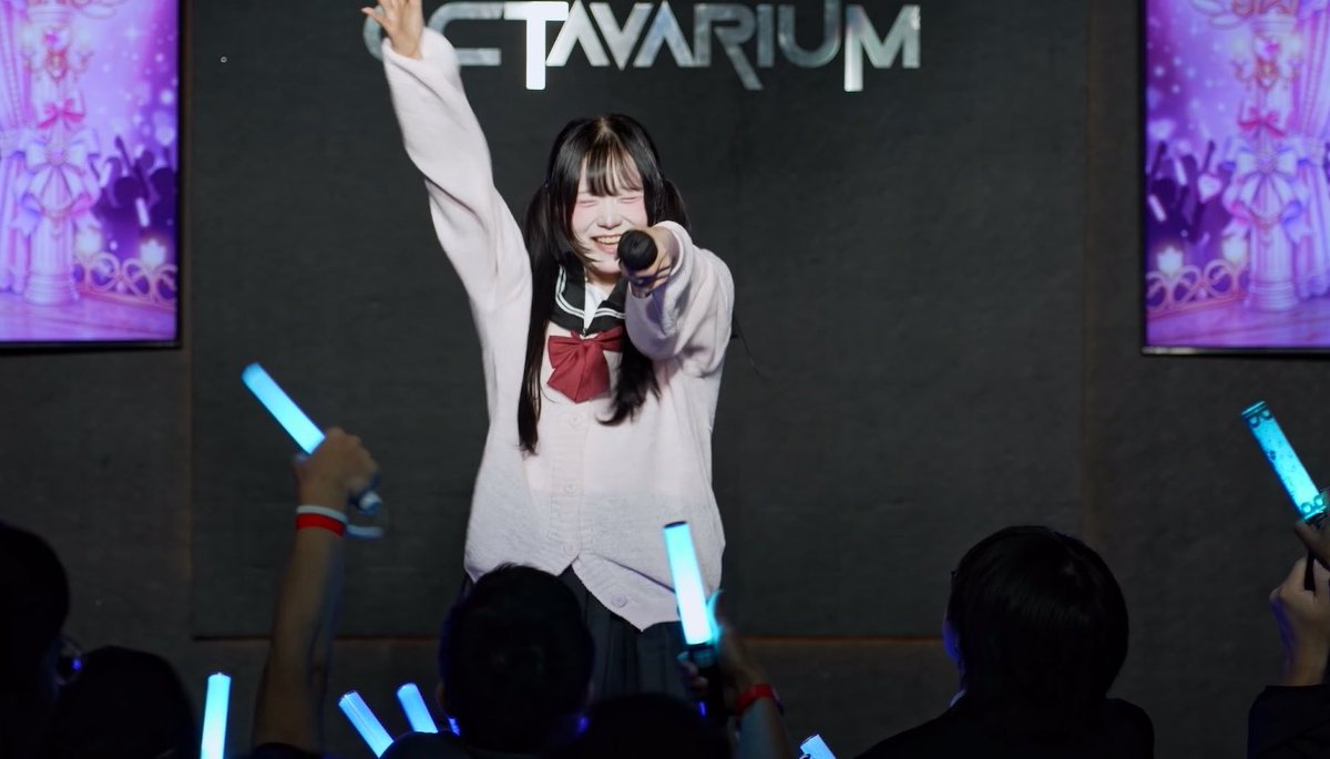 A young woman in a Japanese school uniform (sailor-style with white blouse and red bow) performs on stage at an 'OTAVARIUM' event, holding a microphone and smiling with her arm raised while fans wave blue light sticks.