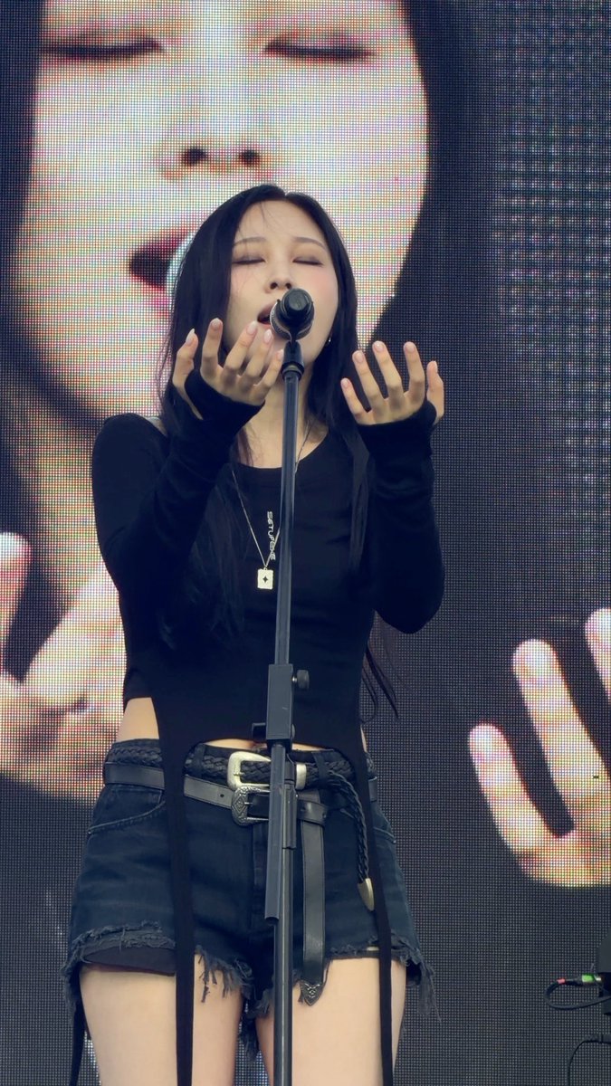 A woman with long dark hair, wearing a black top and distressed denim skirt with a decorative belt, sings into a microphone on stage with her hands raised and eyes closed.