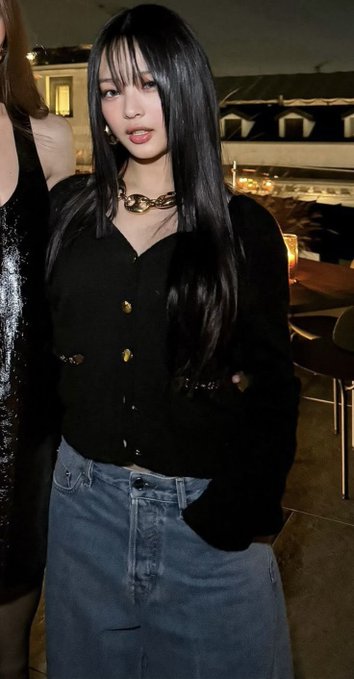 A young woman with long dark hair wearing a black button-up cardigan with gold buttons, a denim skirt, and a chunky gold chain necklace, posing at what appears to be an indoor event or venue.