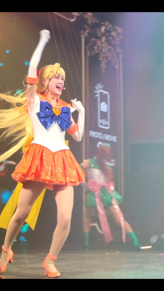 A performer dressed as Sailor Moon (blonde pigtails, white and orange outfit, blue bow) is on stage mid-performance, with another character in green and pink visible in the background.