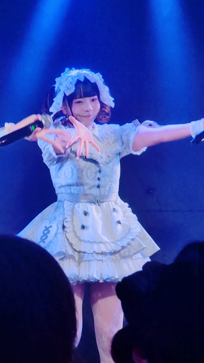 A performer in a frilly light blue maid-style dress with white stockings performing on a stage with blue lighting, reaching toward the camera.