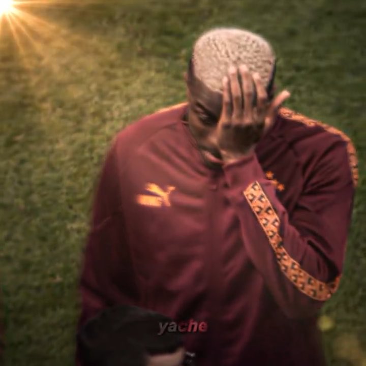 A man in a maroon Puma tracksuit kneels on grass with his hand on his head in a moment of apparent disappointment or exhaustion, with a 'yache' watermark visible.