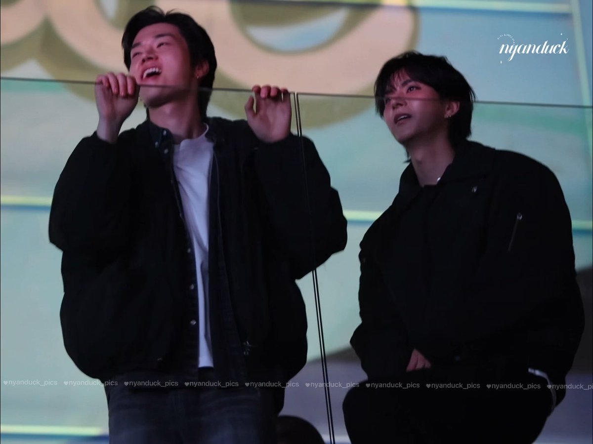 Two young men behind a glass barrier, one laughing joyfully with his hands raised, both wearing black jackets, watermarked with 'nyanduck_pics'.