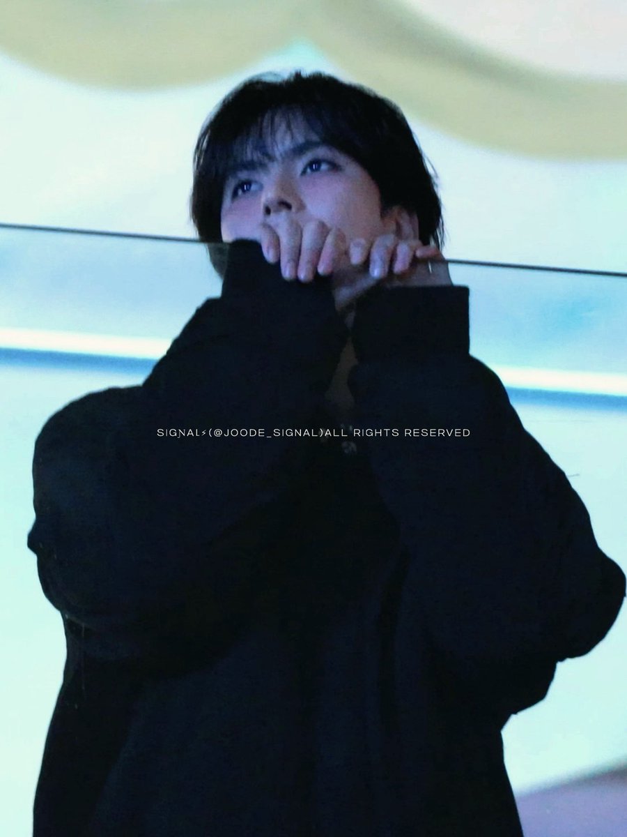 A photo of a young man with dark hair wearing a heavy black coat, resting his chin on his hands in front of a horizontally lit blue background, with a watermark reading 'SIGNAL (@JOODE_SIGNAL)ALL RIGHTS RESERVED'.