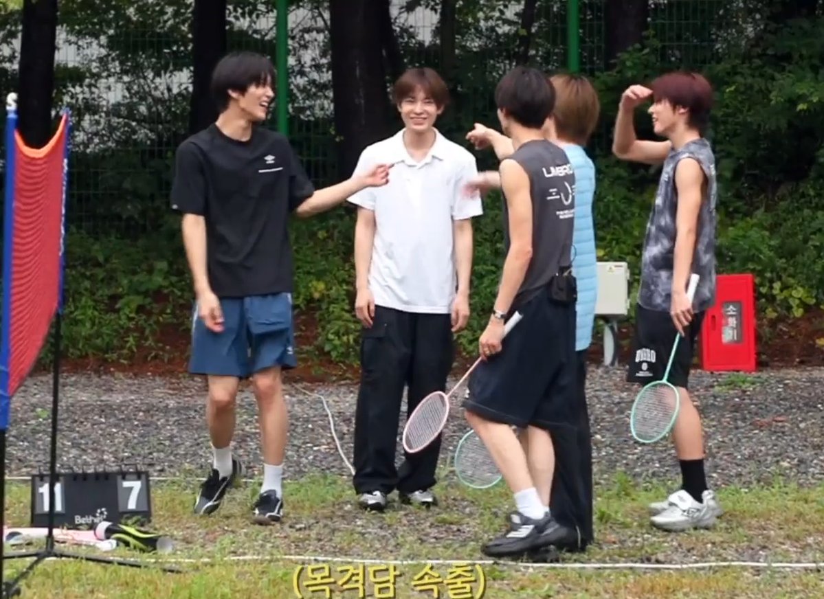 A group of young men, likely a K-pop group, stand together on an outdoor badminton court, with a scoreboard showing "11" and "7" and Korean text "(뭉칫탕 승호)" at the bottom, capturing a lighthearted moment as one member (Saku) reacts to being caught in an embarrassing mishap during their game.