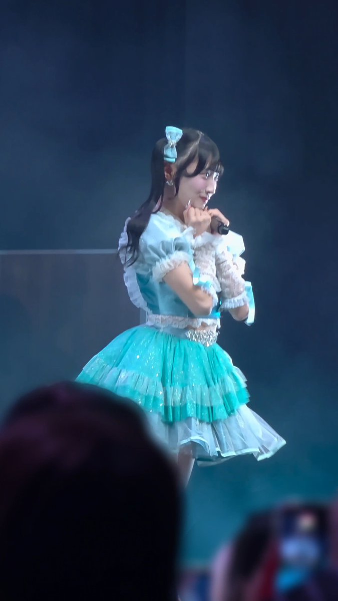 A young woman performs on stage wearing a sparkly teal and white frilled dress with a matching hair bow, holding a microphone with a soft smile.