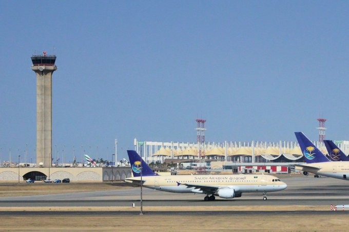 Two Saudi Arabian Airlines passenger jets taxi on a runway, with a large stadium visible in the background alongside airport control towers.