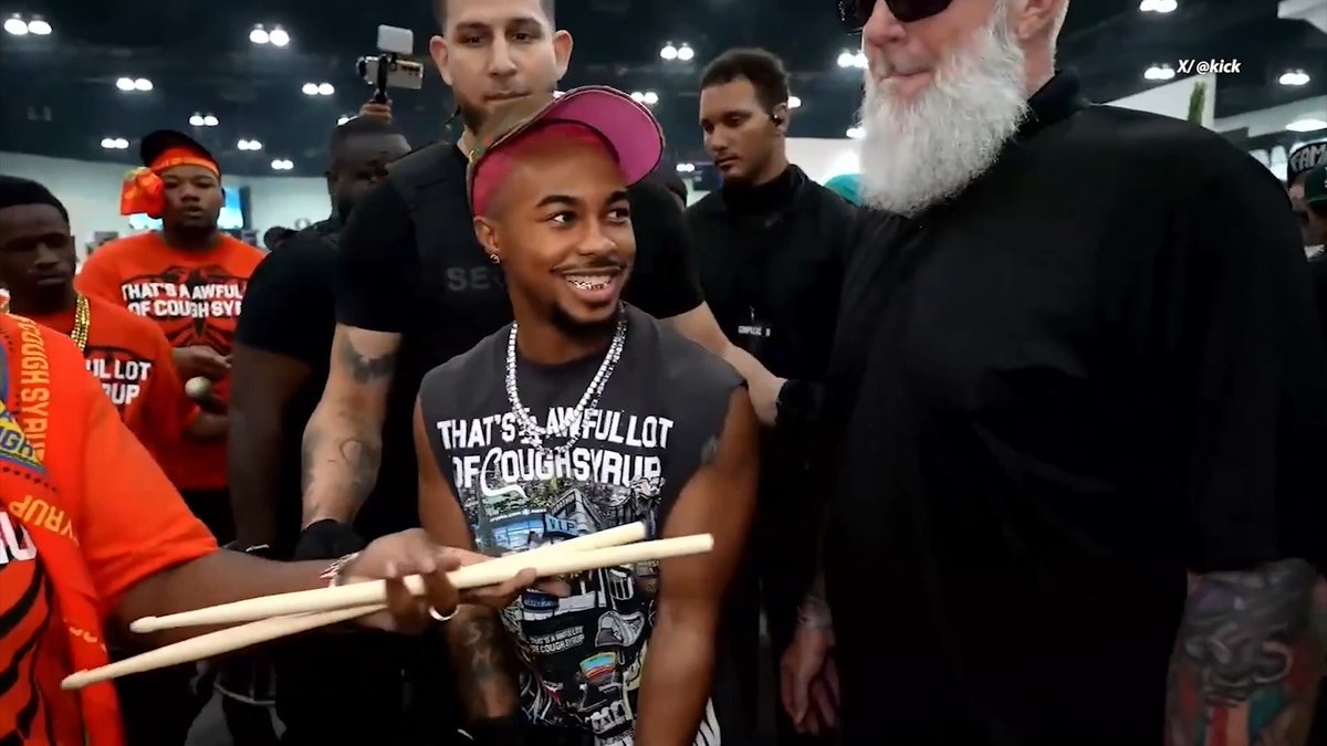 A smiling young man in a pink cap wears a black sleeveless shirt reading "THAT'S A AWFULLOT OF COUGHSYRUP" and holds drumsticks extended toward him by Travis Barker, the bearded, tattooed drummer on the right, during the ComplexCon event referenced in the post.
